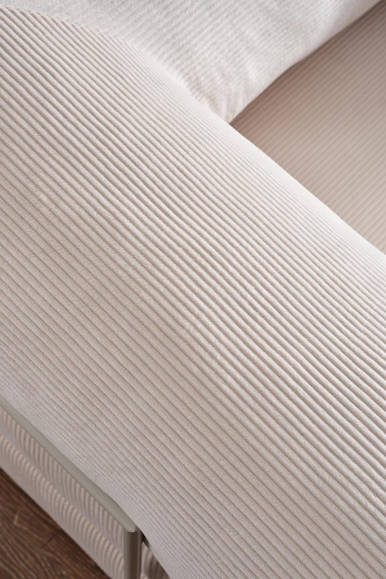 May include: Close-up of a light beige corduroy sofa. The fabric has a ribbed texture, with parallel lines running across the surface. The sofa appears to be part of a larger set, with a portion of another cushion visible.