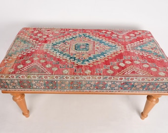 Könnte beinhalten: Ein rechteckiger Hocker mit einer gemusterten Teppichoberfläche in Rot, Türkis und Beige. Der Hocker hat vier hellfarbige Holzbeine. Das Teppichdesign zeigt eine zentrale Raute mit geometrischen Mustern. Der Hocker ist ca. 100 cm lang.