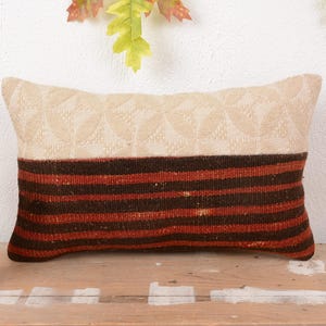 Puede incluir: Cojín decorativo rectangular con diseño bicolor. La parte superior es beige claro con un patrón repetido, mientras que la inferior presenta rayas horizontales en tonos marrón y rojo. Descansa sobre una superficie de madera. Mide unos 40 cm de largo.