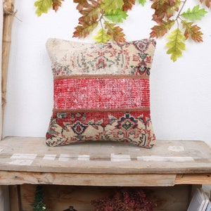 May include: Square decorative pillow with a patchwork design. The pillow features a central red section, bordered by sections with a beige and floral pattern. The pillow is placed on a wooden table.