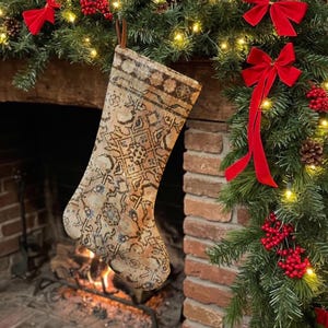 Puede incluir: Un calcetín navideño con un patrón de alfombra oriental intrincado y desteñido en tonos beige, marrón y azul apagado. El calcetín tiene un lazo de cuero marrón para colgar en la parte superior y está hecho de un material de tela tejida.