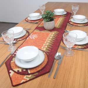 Puede incluir: Un camino de mesa con un diseño geométrico en rojo, naranja y verde. El camino de mesa está colocado sobre una mesa de madera con platos, cuencos y vasos blancos.