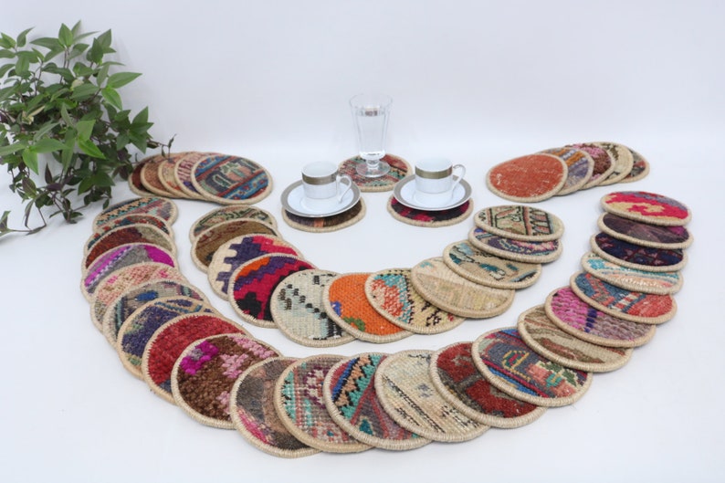 May include: A set of 20 colorful round coasters made from woven fabric with a jute trim. The coasters are arranged in a semi-circle pattern on a white surface. Two white porcelain teacups and saucers are placed on two of the coasters.