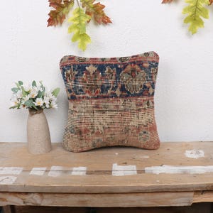 May include: A square throw pillow with a colorful, vintage-style patterned fabric. The pillow has a dark blue border with red, orange, and yellow accents. The fabric is woven with a variety of textures and patterns.