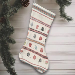 May include: Cream-colored Christmas stocking with a geometric pattern in red, black, blue, and green. The stocking is hanging from a silver loop against a white wooden background, with a pine branch and ornaments in the background.