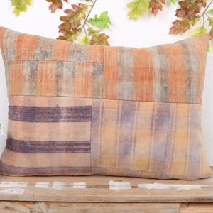 May include: Rectangular decorative pillow with a patchwork design. The pillow features a combination of orange, purple, and yellow hues. The top section has vertical stripes, while the bottom has horizontal stripes and a plaid pattern. The pillow is placed on a wooden surface.
