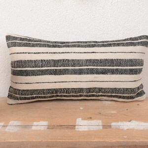 Puede incluir: Cojín decorativo rectangular con rayas horizontales negras sobre fondo crema. El cojín está hecho de tela texturizada y descansa sobre una superficie de madera. Un artículo decorativo para cama o sofá.