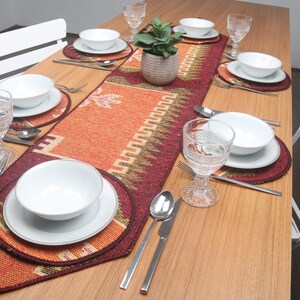Puede incluir: Una mesa de madera puesta con un camino de mesa estampado en rojo, naranja y verde. Se colocan cuencos y platos de cerámica blanca encima del camino de mesa, junto con cubiertos de plata y copas de vino de vidrio transparente.
