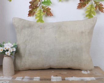 May include: A rectangular, light beige pillow with a textured surface. The pillow is placed on a wooden surface, with a small vase of white flowers to the left. Decorative leaves are above.