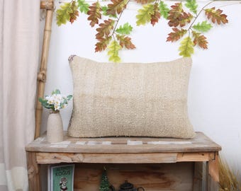 May include: A rectangular beige pillow with a textured surface rests on a wooden table. The pillow has a neutral color and a woven appearance. Decorative leaves hang above the pillow, adding a natural touch to the scene.