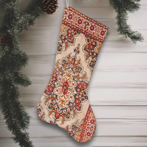 May include: A Christmas stocking with a cream-colored background and a detailed floral pattern in red, orange, blue, and gold. The stocking is hanging from a silver loop against a white wooden background, with a pine branch and ornaments in the corner.