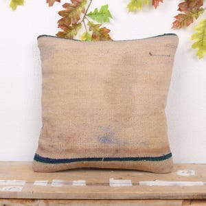 May include: A square, beige pillow with a textured surface. The pillow has a dark blue border and a few faded blue and red marks. It rests on a wooden surface, with autumn leaves in the background.