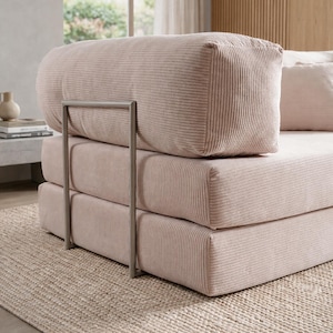 May include: A light pink corduroy sofa with a metal frame. The sofa has a stacked design with a rounded backrest and a rectangular seat. The sofa is placed on a beige woven rug, creating a cozy and inviting atmosphere.