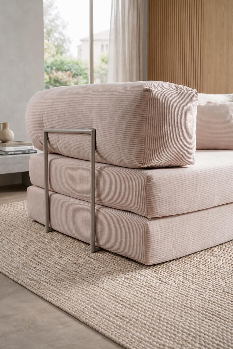 May include: A light pink corduroy sofa with a metal frame. The sofa has a stacked design with a rounded backrest and a rectangular seat. The sofa is placed on a beige woven rug, creating a cozy and inviting atmosphere.