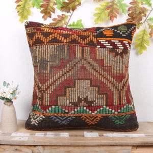 May include: Square decorative pillow with a geometric pattern in shades of red, brown, and orange. The pillow has a dark brown base and a mix of textures. A small vase with white flowers sits nearby.