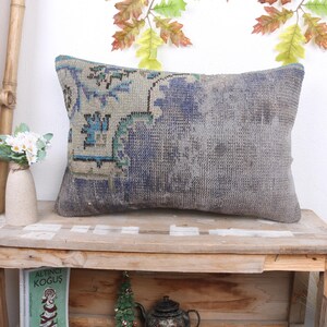 May include: Rectangular decorative pillow with a faded, distressed appearance. The pillow features a central design in shades of blue and teal on a gray background. The pillow is placed on a rustic wooden table, suggesting a vintage or bohemian style.