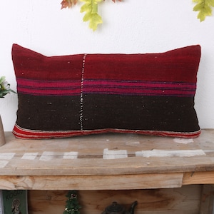 Puede incluir: Una almohada rectangular de color rojo, marrón y rosa a rayas con una textura tejida. La almohada es larga y estrecha.