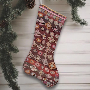 May include: A Christmas stocking with a patterned design in shades of burgundy, pink, orange, and white. The stocking is hanging from a silver loop, with a Christmas tree branch in the background.