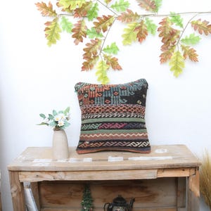 May include: A decorative throw pillow with a colorful geometric pattern in shades of green, blue, black, and orange. The pillow is sitting on a wooden shelf with a vase of white flowers and a potted plant.