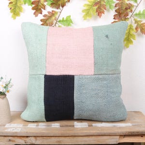 May include: A square decorative pillow with a patchwork design. The pillow features a light green, pink, and navy blue color scheme. It is placed on a wooden table, with a small vase of flowers and decorative leaves in the background.