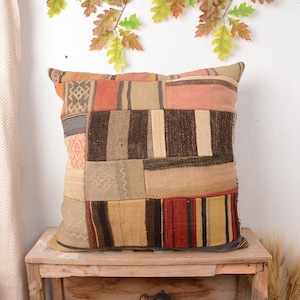 May include: A square patchwork cushion with a variety of colours and textures. The cushion features rectangular and square patches in shades of orange, brown, beige, and red. The cushion is placed on a wooden table.