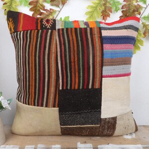 May include: A square patchwork cushion with a variety of colours and patterns. The cushion features vertical stripes, solid blocks, and geometric designs in shades of red, brown, black, and beige. The cushion is approximately 46 cm square.