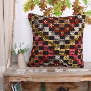 May include: A square decorative pillow with a woven, checkered pattern in black, red, yellow, and white. The pillow is placed on a rustic wooden table, with a vase of white flowers and autumn leaves in the background.