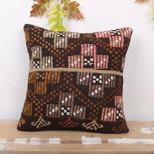 Peut inclure: Un coussin carré décoratif avec un motif géométrique dans les tons de marron, rose, orange et blanc. Le coussin est posé sur une surface en bois, avec des feuilles d'automne en arrière-plan. Le design du coussin comporte une bande centrale.