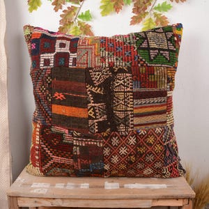 Op de afbeelding: Een vierkant patchwork kussen met diverse geometrische patronen in rood, bruin, oranje en groen. Het kussen is gemaakt van een geweven stof en ligt op een houten oppervlak.