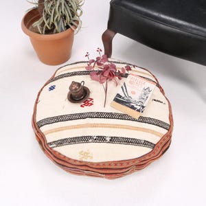 May include: Round, cream-colored floor cushion with a striped pattern in black, tan, and red. A small metal cup and saucer, a sprig of flowers, and a book are on top. A potted plant and a black chair are in the background.