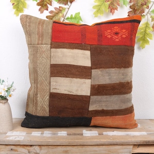 May include: A square, patchwork pillow with a variety of brown, beige, and orange fabrics. The pillow has a geometric design and is placed on a wooden surface. Decorative leaves are in the background.