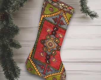 May include: A festive Christmas stocking with a colourful, patterned design. The stocking has a red central field with a detailed floral motif, bordered by green and yellow geometric patterns. It hangs from a white string against a wooden background, evoking a holiday theme.