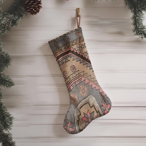 Op de afbeelding: Een kerstkous met een geweven patroon in grijstinten, beige en gedempt roze. De kous heeft een geometrisch ontwerp met herhalende bloemachtige motieven en een ophanglus aan de bovenkant, hangend aan een haakje aan een witte muur.