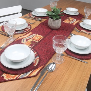 Puede incluir: Una mesa de madera puesta con un mantel individual rojo y dorado, platos, cuencos y vasos blancos. La mesa está puesta para una cena formal con cubiertos y una pequeña planta en maceta en el centro.