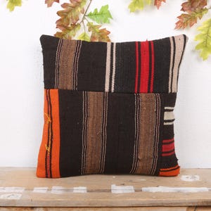 Puede incluir: Un cojín cuadrado con un patrón de rayas en negro, marrón, rojo, naranja y blanco. El cojín está hecho de una tela tejida y se encuentra sobre una superficie de madera. Hay hojas decorativas en el fondo.