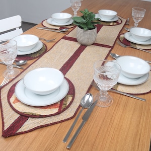 May include: A wooden table set with a brown and red table runner, white plates, bowls, and glasses. The table is set for a dinner party with silverware and a small potted plant in the center.