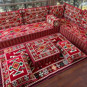 May include: A red and white patterned floor cushion set with intricate geometric designs. The set includes a large sofa-like cushion, smaller square cushions, and a rectangular rug. The cushions are arranged in a traditional Moroccan style.