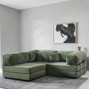May include: A large, olive green sectional sofa with a corduroy texture. The sofa has a modular design with multiple cushions and pillows. The room features a gray rug and a framed abstract painting on the wall.
