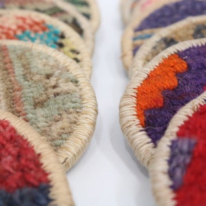 May include: A stack of colorful round woven coasters with a beige trim. The coasters are made of a soft, textured fabric and feature geometric patterns in shades of red, orange, purple, blue, and green.