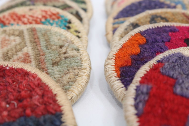 May include: A stack of colorful round woven coasters with a beige trim. The coasters are made of a soft, textured fabric and feature geometric patterns in shades of red, orange, purple, blue, and green.