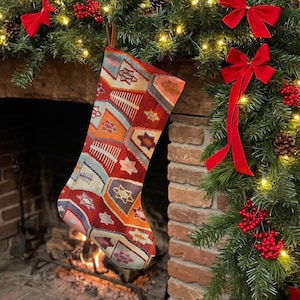 Könnte beinhalten: Ein Weihnachtsstrumpf mit einem mehrfarbigen geometrischen Muster in Rot-, Orange-, Blau- und Cremetönen. Der Strumpf hat eine braune Aufhängeschlaufe aus Leder an der Oberseite und besteht aus einem gewebten Stoff mit einem sich wiederholenden Muster.