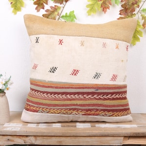 May include: A decorative pillow with a beige top and bottom, and a cream-colored center section. The center section has small red and black geometric patterns. The bottom section has a horizontal band of colorful stripes. The pillow is on a wooden surface.