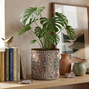 Può includere: Un coprivaso cilindrico in tessuto con un dettagliato motivo geometrico nei toni del beige, blu e ruggine. Il coprivaso contiene una pianta di monstera verde con grandi foglie intagliate. Il bordo superiore è rifinito in marrone scuro.