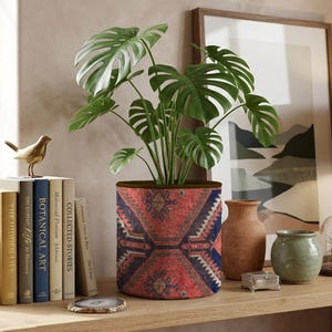 Puede incluir: Una funda cilíndrica de tela para macetas con un patrón geométrico rojo y azul y un borde marrón oscuro. Una planta Monstera verde con hojas grandes y divididas está colocada dentro del recipiente, sobre una superficie de madera.