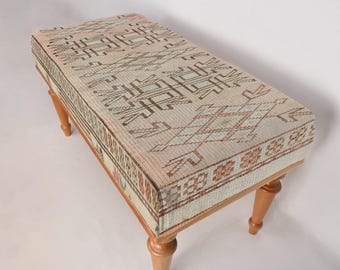 May include: A wooden bench with a patterned cushion. The cushion is a light pink and white with a geometric design. The bench has four legs and is made of wood.