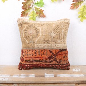 May include: Square decorative pillow with a beige and orange patterned design. The top half of the pillow is beige with a geometric pattern, while the bottom half is orange with a darker, more intricate design. The pillow is placed on a wooden surface.