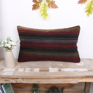 May include: A long rectangular pillow with a striped pattern in shades of red, black, brown, and gray. The pillow is made of woven fabric and has a rustic, handmade look.