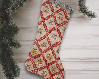 May include: A festive Christmas stocking featuring a beige background with a red and blue geometric design. The stocking is adorned with floral patterns in yellow and blue. A white ribbon is attached for hanging.