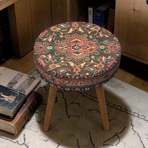 Può includere: Uno sgabello rotondo con un piano fantasia con un motivo floreale centrale nei toni del rosso, rosa e blu su sfondo marrone. Lo sgabello ha tre gambe in legno. Il design generale ricorda un tappeto vintage.