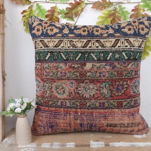 May include: A square cushion with a patchwork design in shades of blue, green, red, and beige. The cushion has a woven fabric with a vintage or antique appearance. The cushion is a decorative item.
